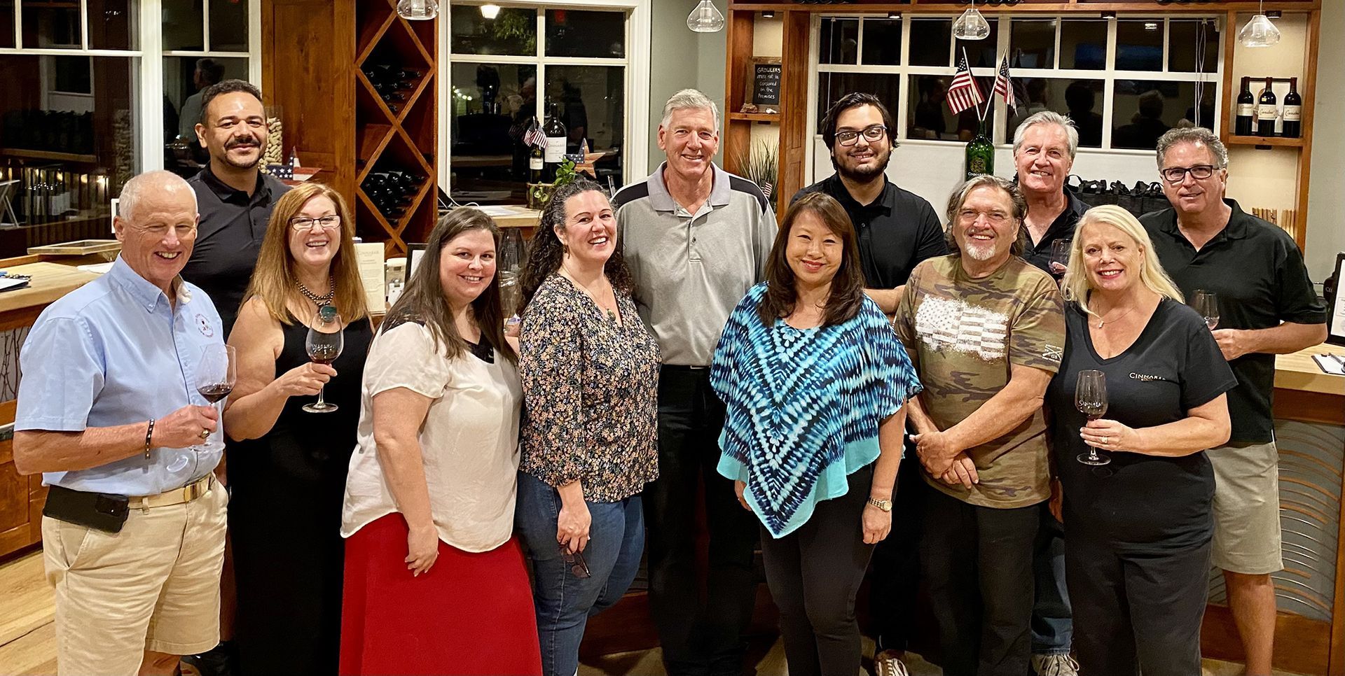 A group of 12 people smiling together while holding wine glasses inside a winery or tasting room.