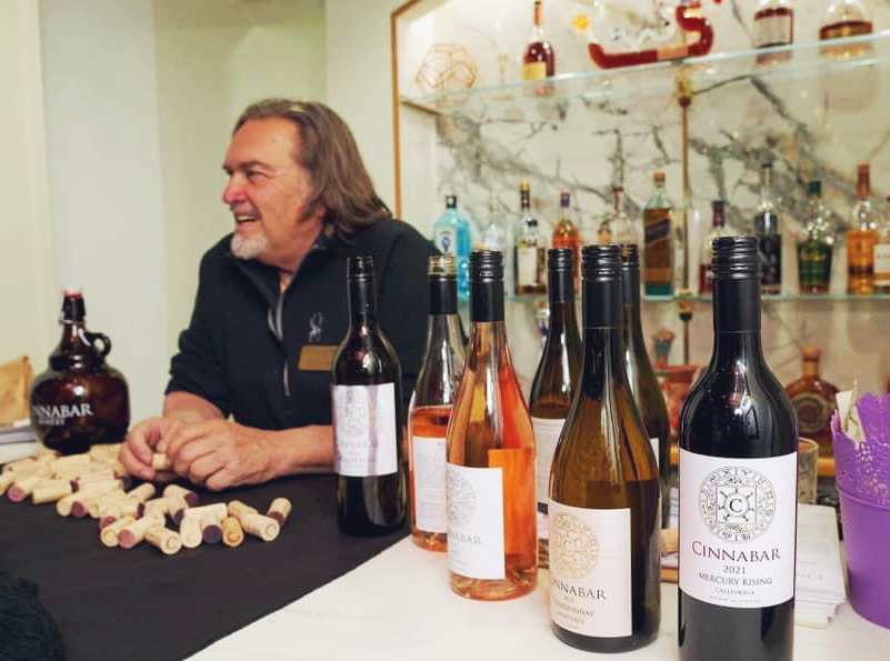 A person sits behind a table filled with wine bottles and corks in a room with a back bar displaying spirits.