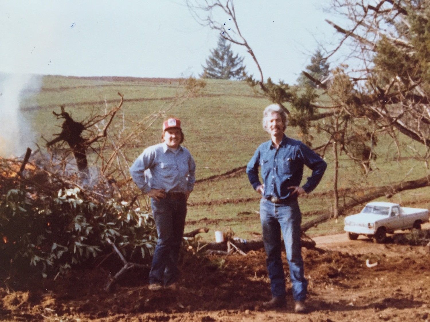 Two people stand in a field next to a burning brush pile and a white pickup truck, with rolling hills in the background.