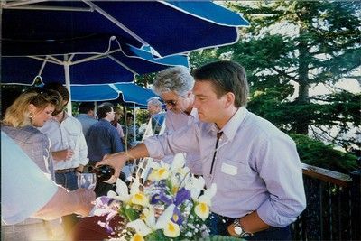 People socialize under blue umbrellas on a patio; a man pours wine into a glass near a vase of flowers.