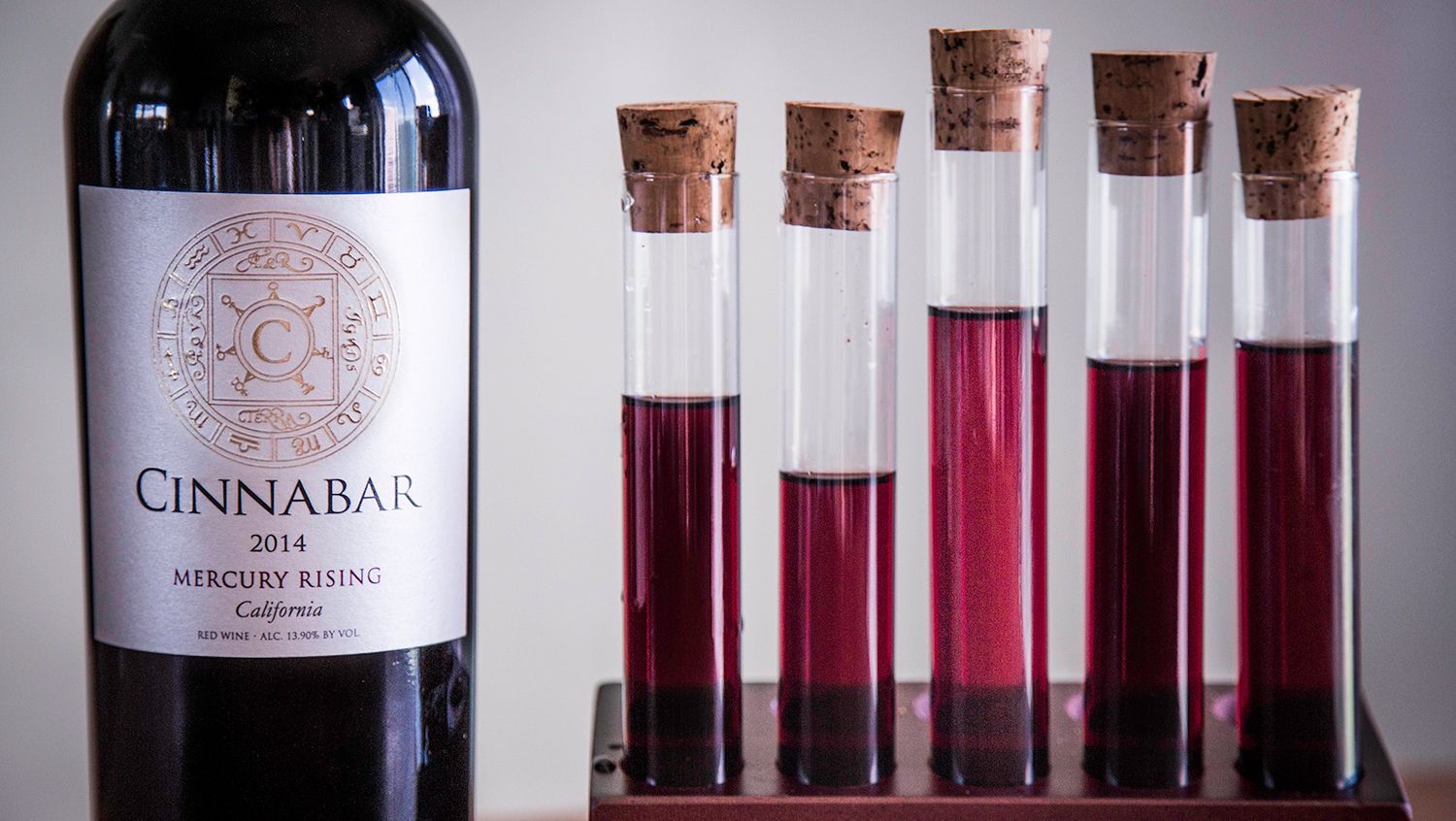 A bottle of Cinnabar 2013 red wine next to a rack of five corked test tubes filled with varying amounts of red wine.