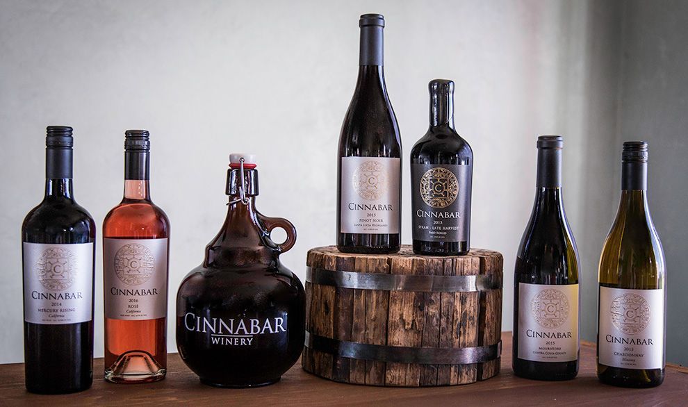 A row of Cinnabar Winery bottles and a jug displayed on a wooden surface, with a small barrel under two of the bottles.