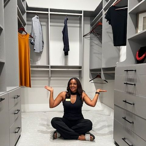 Woman in black outfit sits in a gray walk-in closet, arms outstretched.