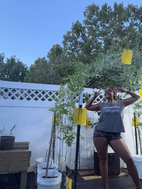 Woman in a garden, arms raised, smiling. Green plants in containers, white fence, blue sky.