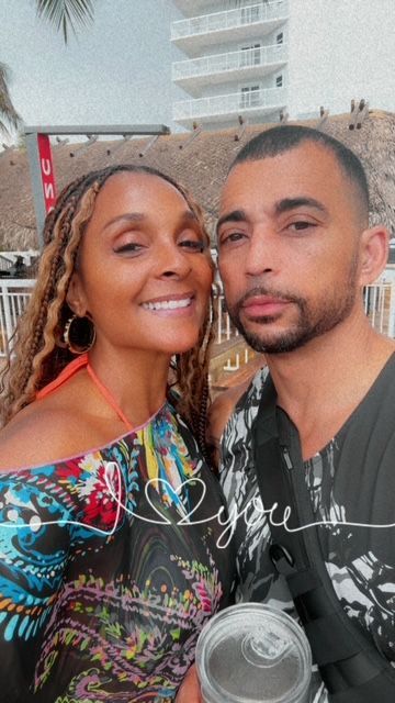 Woman and man smiling, close-up selfie. Outdoors, sunny. Woman in colorful shawl, man in black shirt.