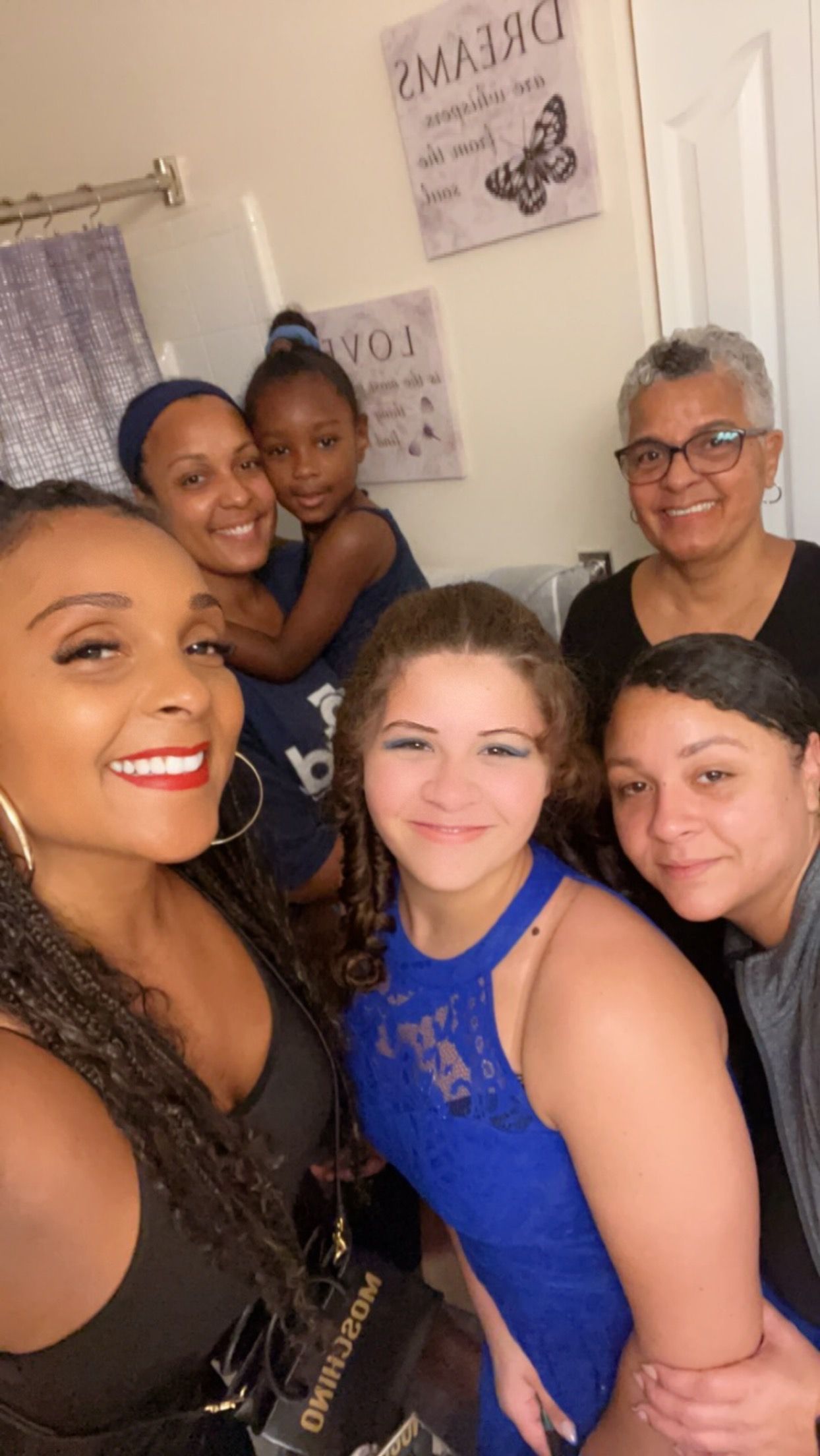 Group of six people smiling in a bathroom; a woman takes a selfie; the rest are behind her.