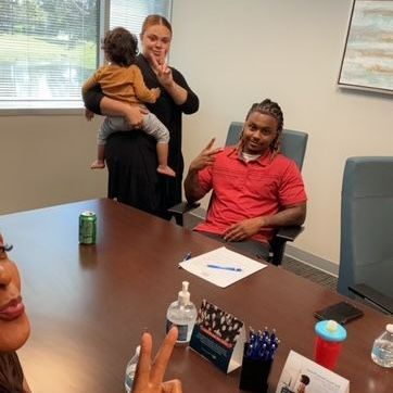 Family in office, signing papers. Man in red shirt, woman holding child, all giving peace signs.