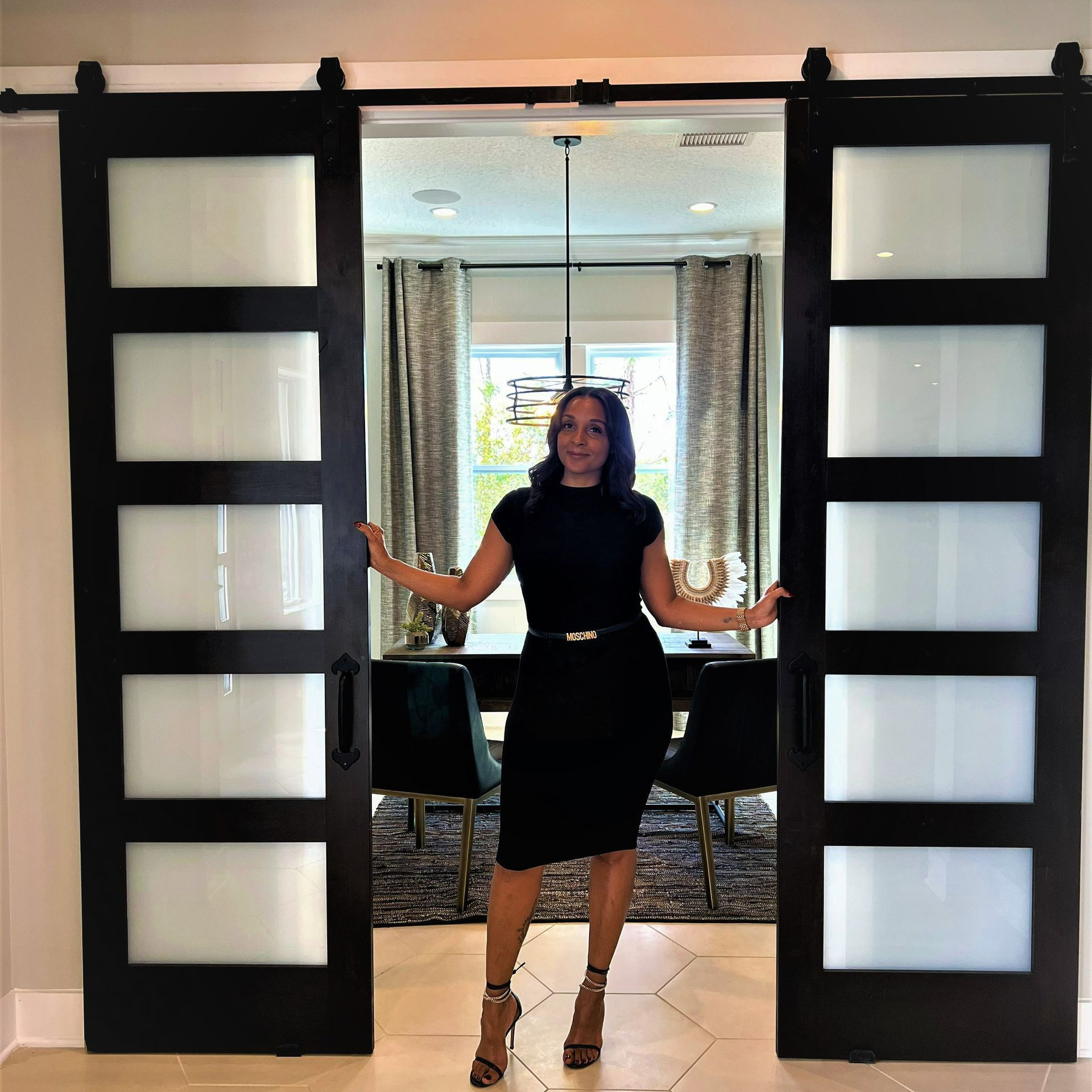 Woman in black dress stands between black sliding glass doors, hands on doors, dining room behind.