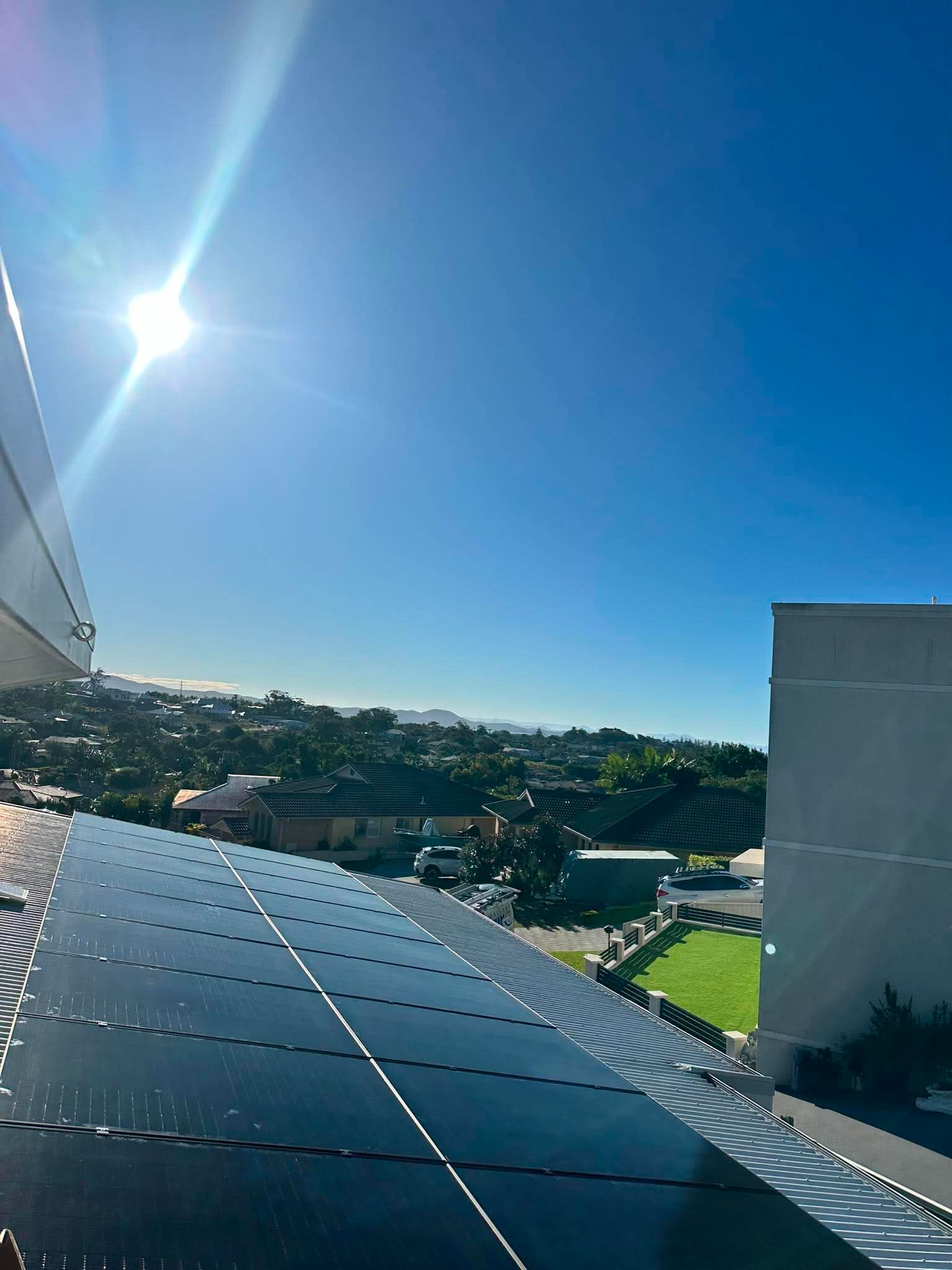 Solar panels on a rooftop under bright sun, overlooking a green hillside and blue sky