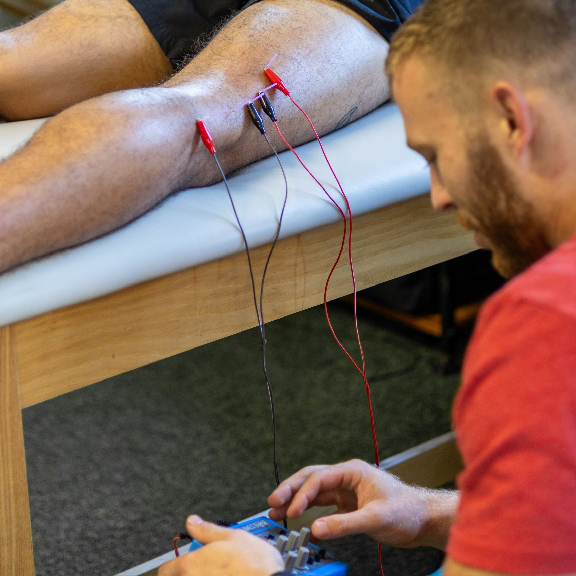 A man receives electrotherapy on his leg from a therapist wearing red shirt indoors.