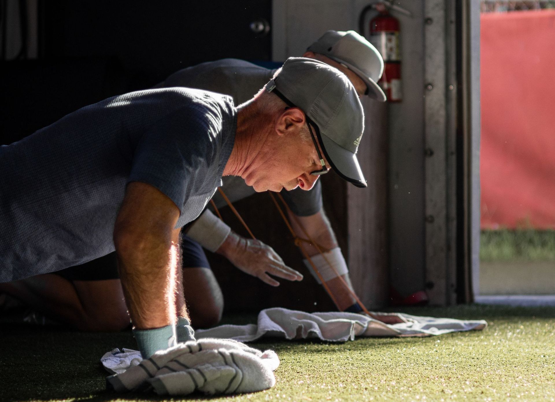 Two men in caps doing push-ups on a green surface; one has a towel nearby.
