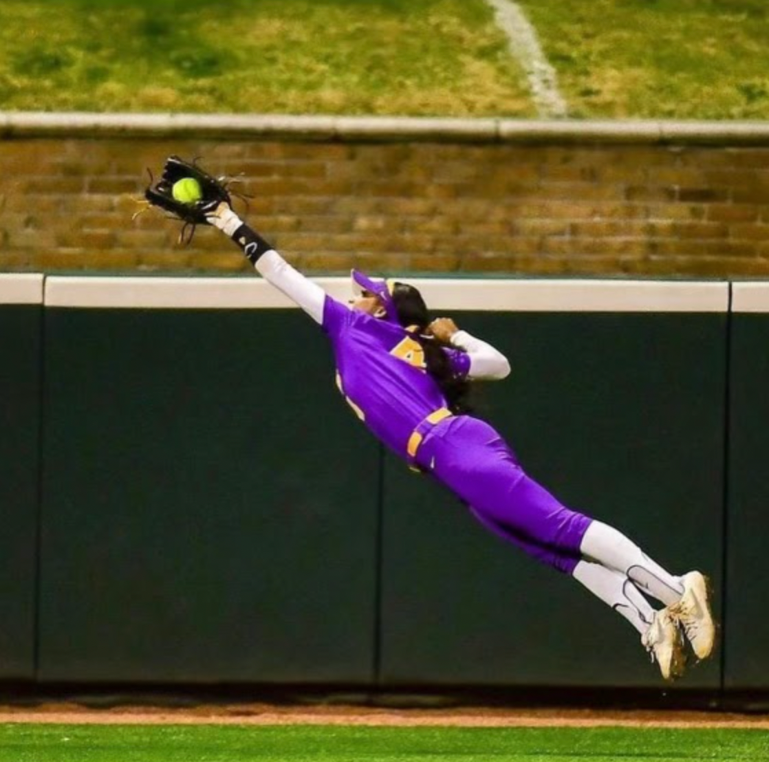 Softball pitcher in maroon jersey, white pants, and black cleats throwing a yellow ball on a dirt field.