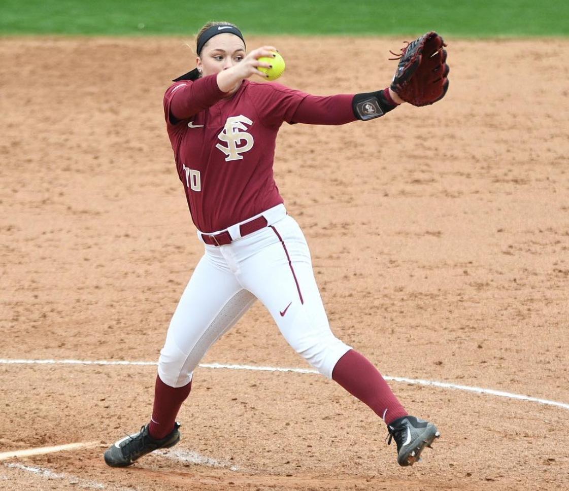 Softball pitcher in maroon jersey, white pants, and black cleats throwing a yellow ball on a dirt field.