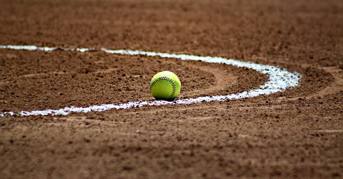 A yellow softball on dirt, near a white foul line curve on a softball field.