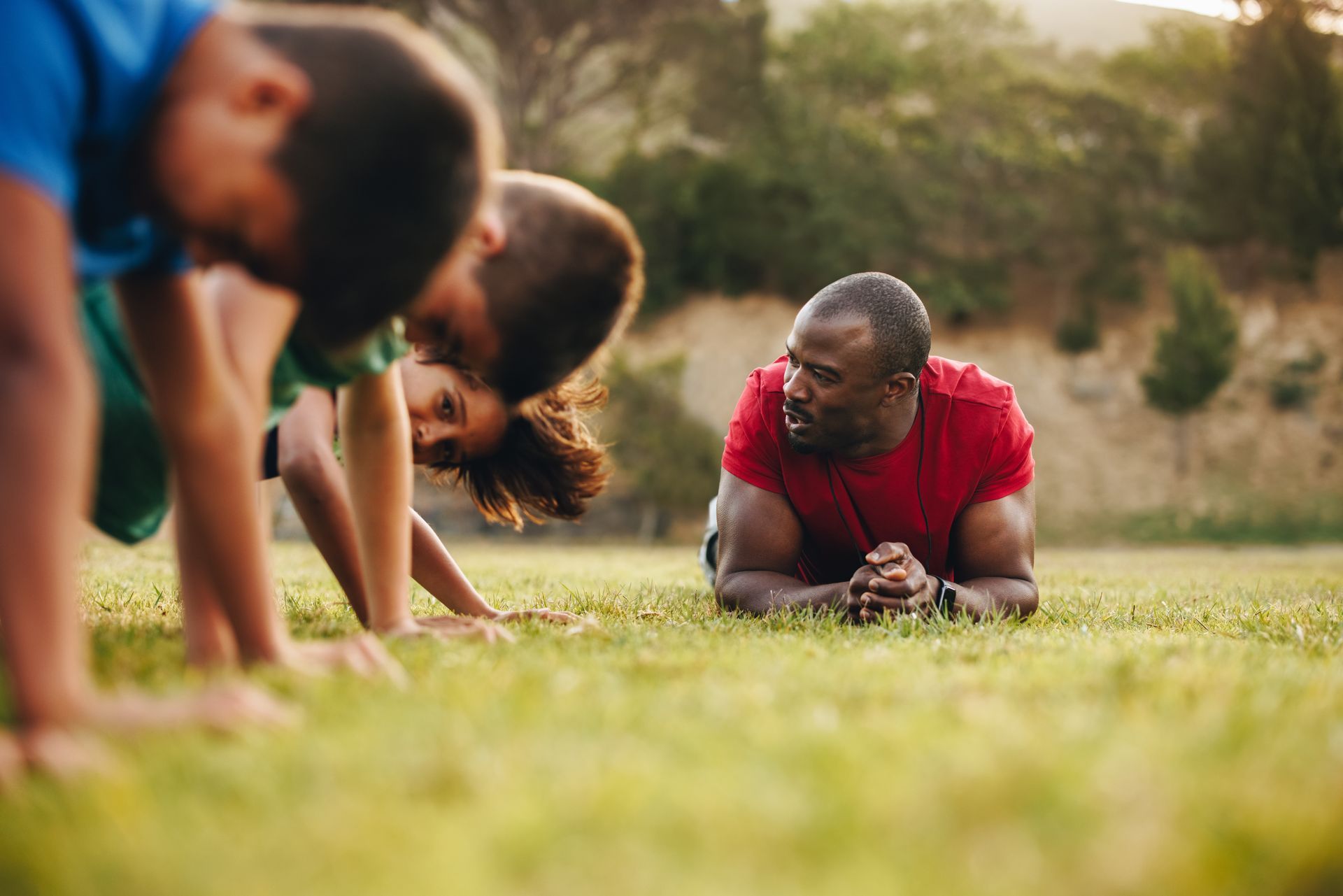 Man supervises children doing push-ups on a grassy field in a park.