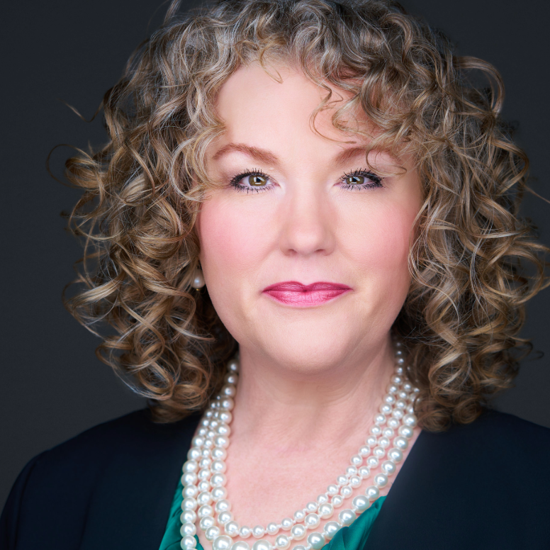 Portrait of a smiling person with curly hair, pearl necklaces, and a dark background