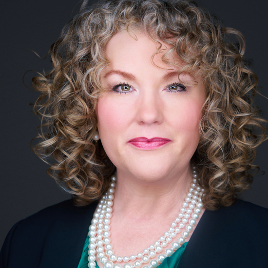 Portrait of a smiling person with curly hair, pearl necklaces, and a dark background