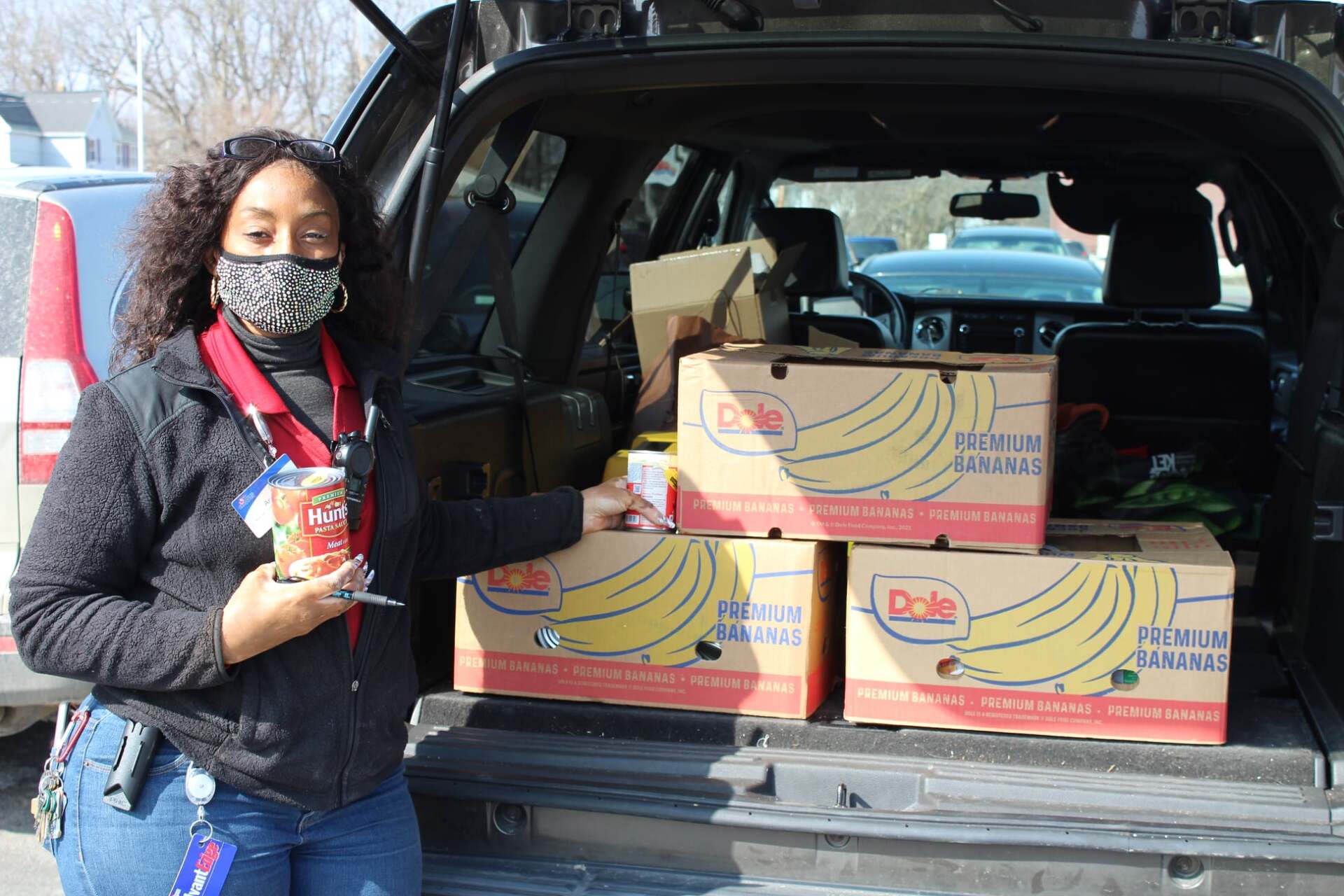 A woman wearing a mask is standing in front of a car filled with banana boxes.