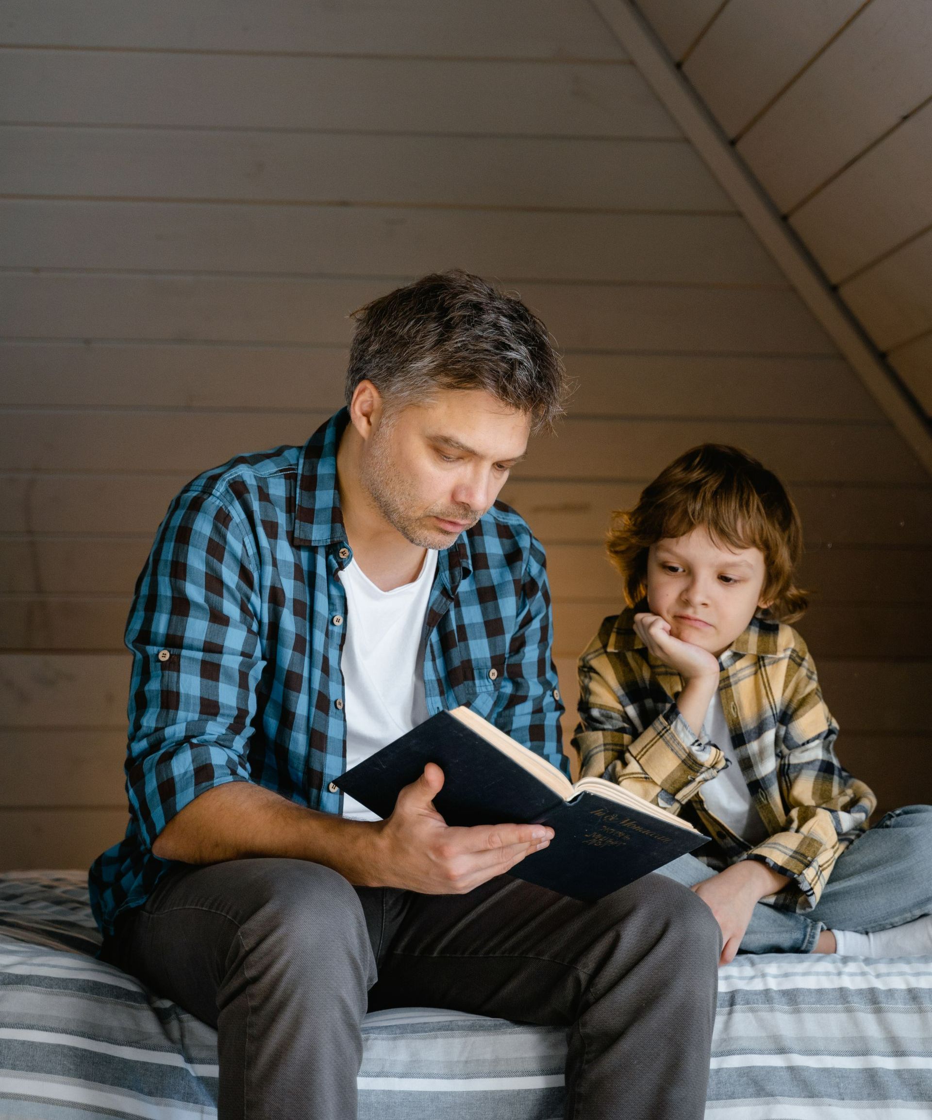 A man and a boy are sitting on a bed reading a book.