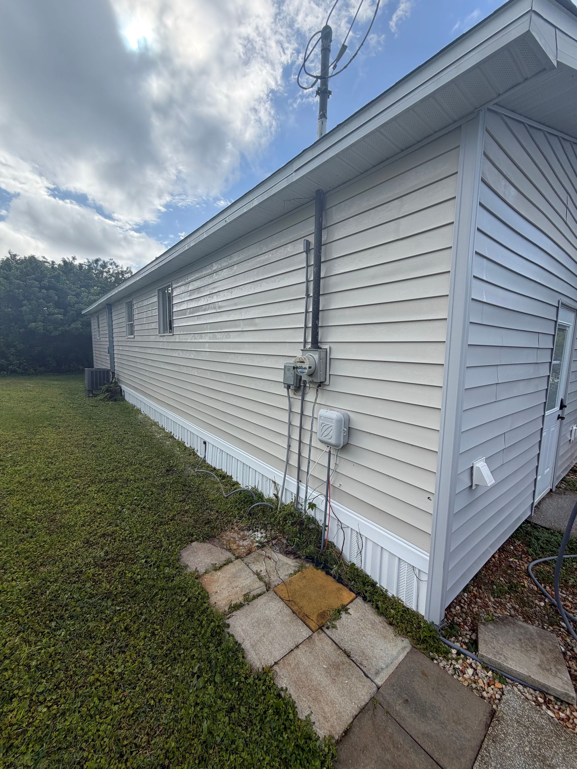 White vinyl house side with utility meter, downspout, and a small stone path beside a grassy yard under a cloudy sky