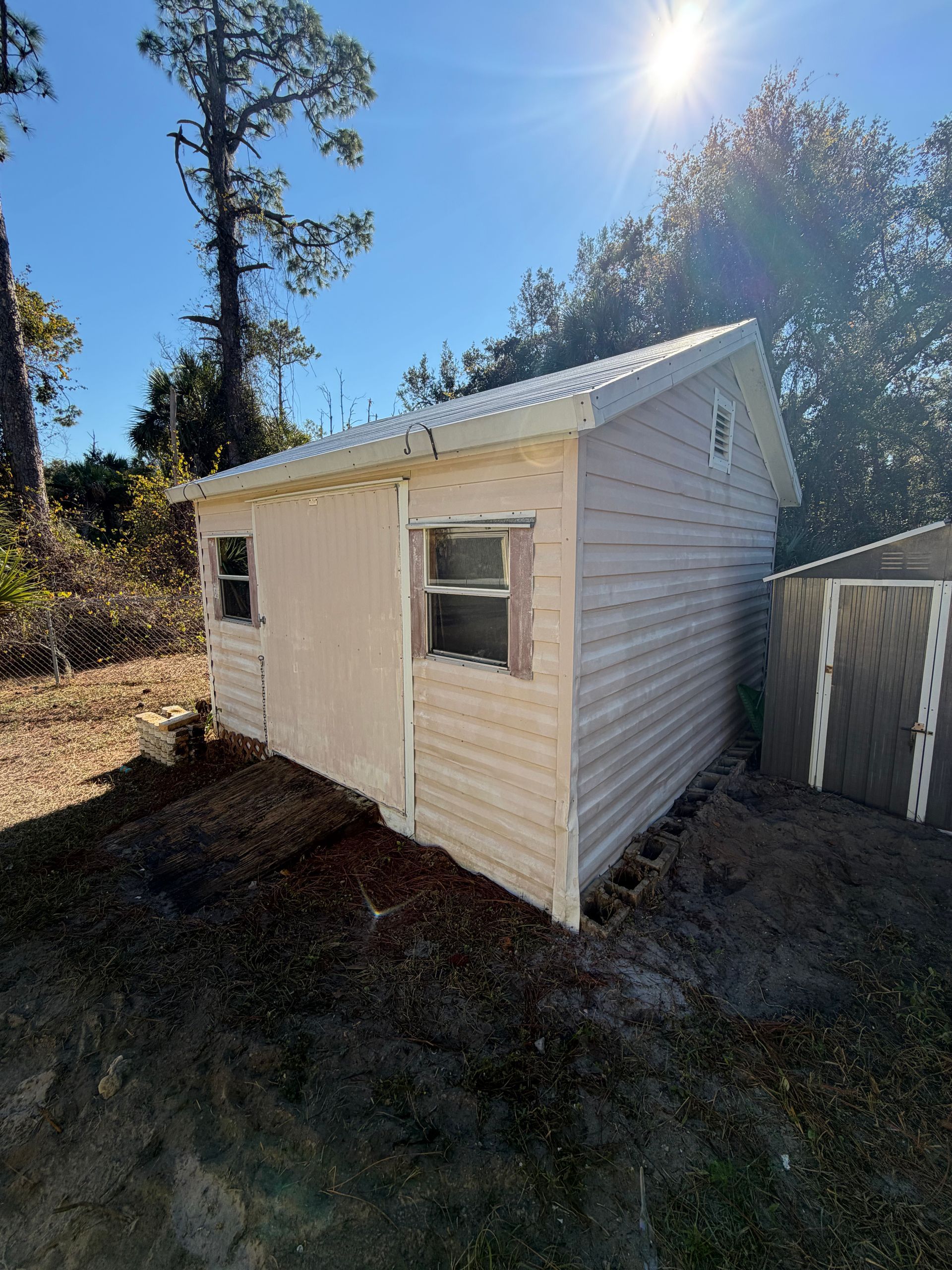 Small beige shed with white trim on a sunny hillside, beside a fence and trees.