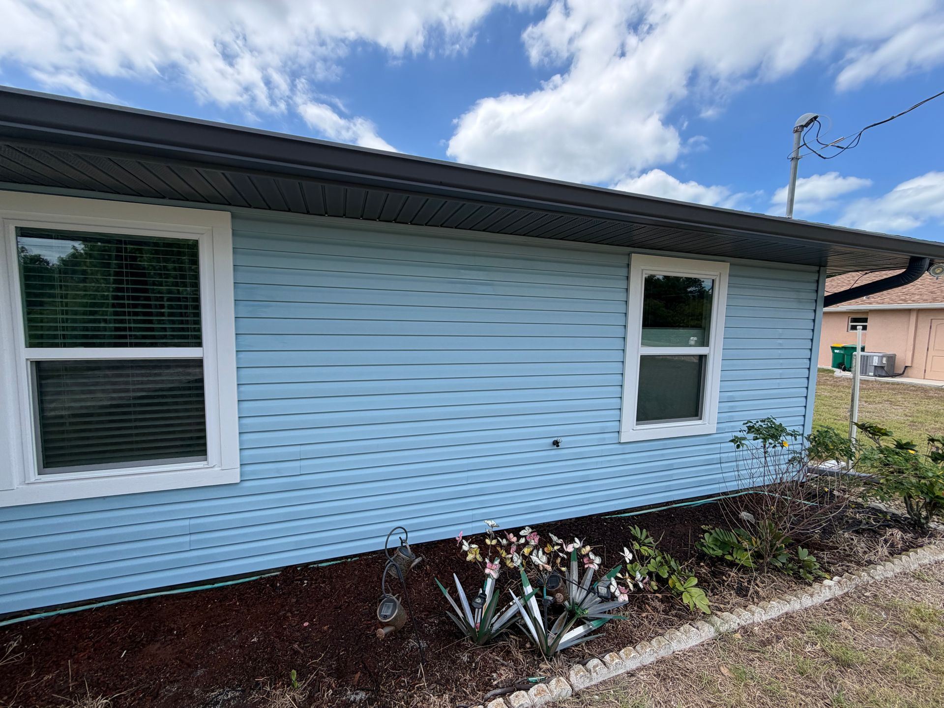 Light blue house exterior with two windows, dark roof, and small shrubs along the foundation