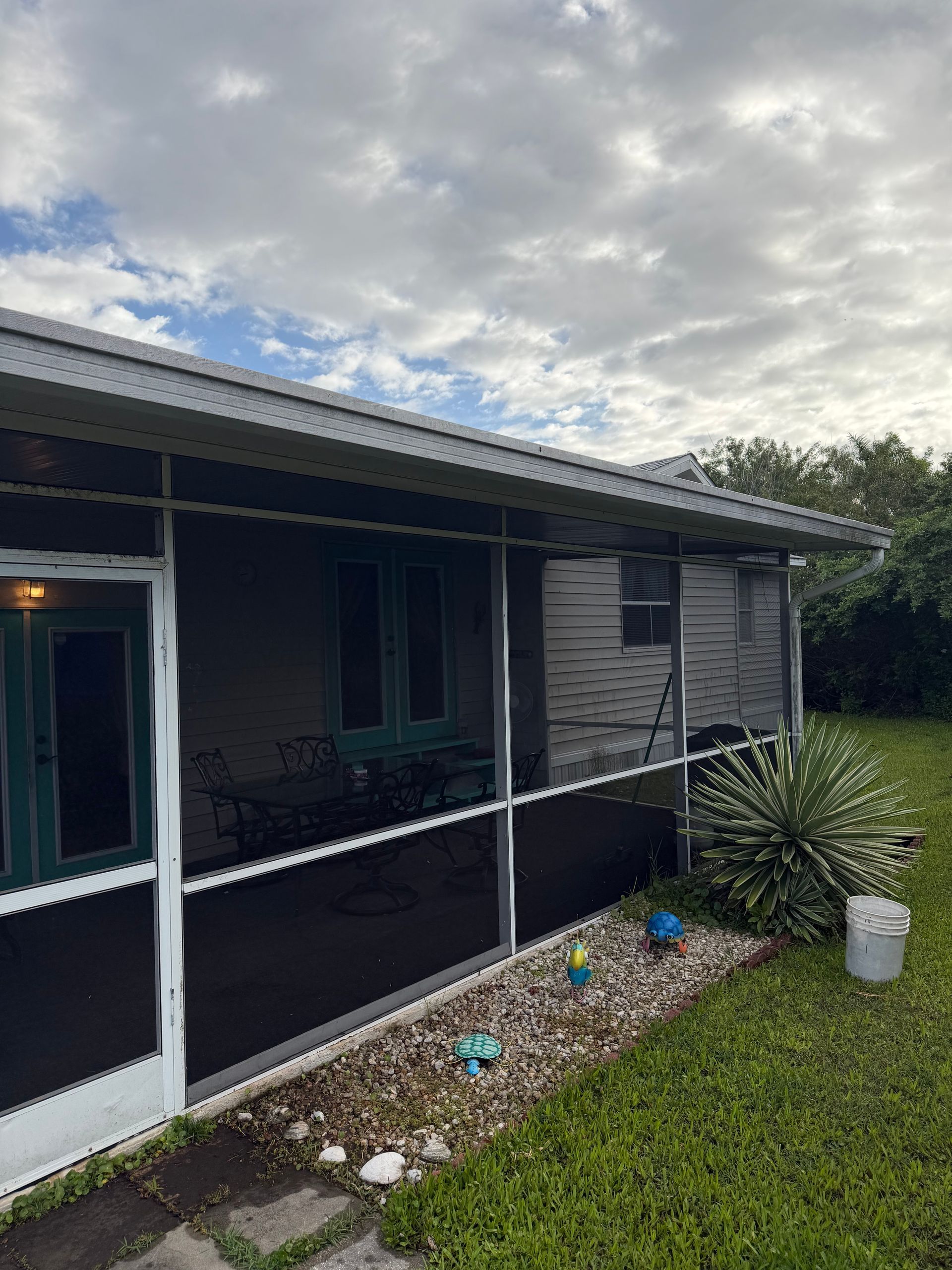 Screened porch of a dark gray house with white trim, black railing, flower bed, and cloudy sky.