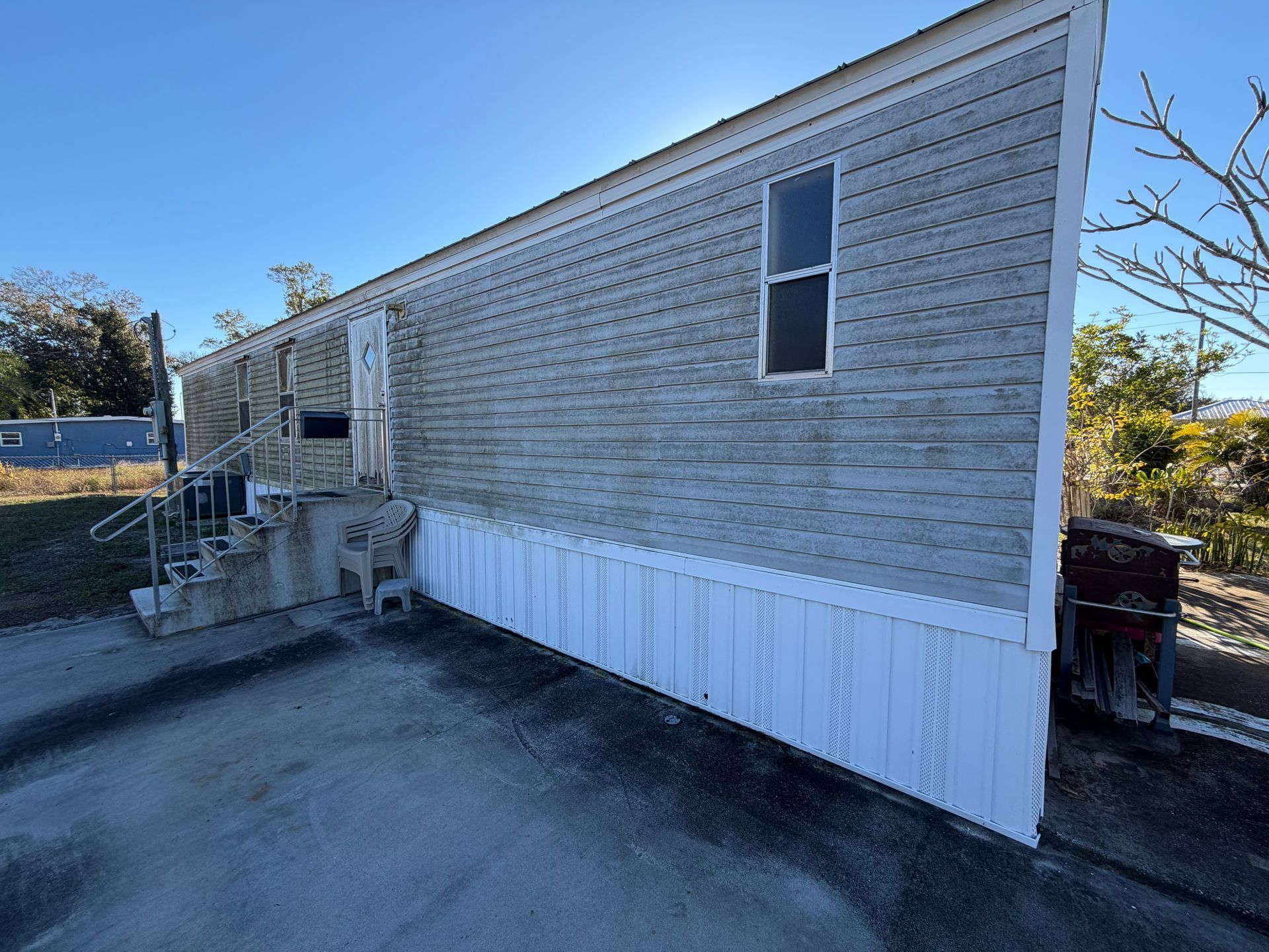 Side view of a weathered mobile home with white skirting, concrete steps, and a paved lot under a clear sky
