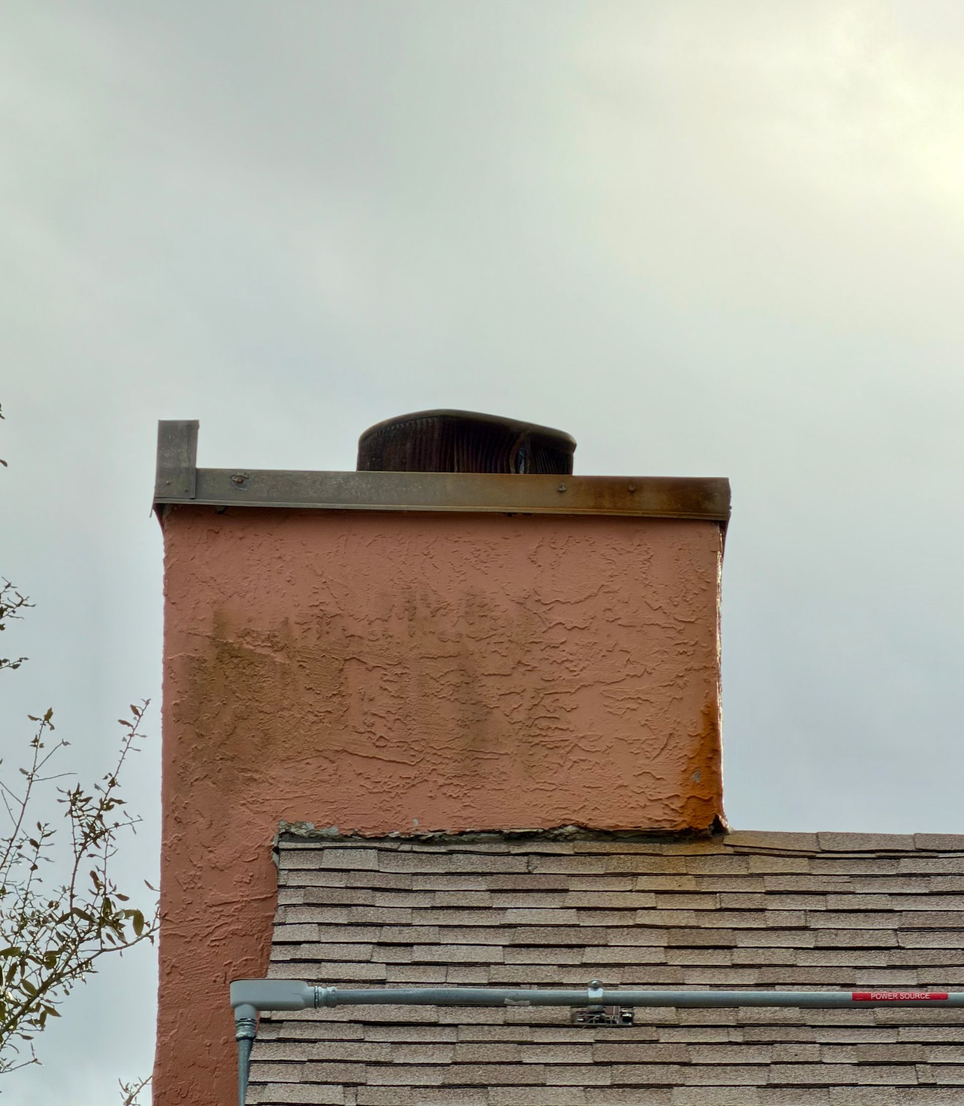Pink stucco chimney with a dark vent, above a gray shingled roof against cloudy sky