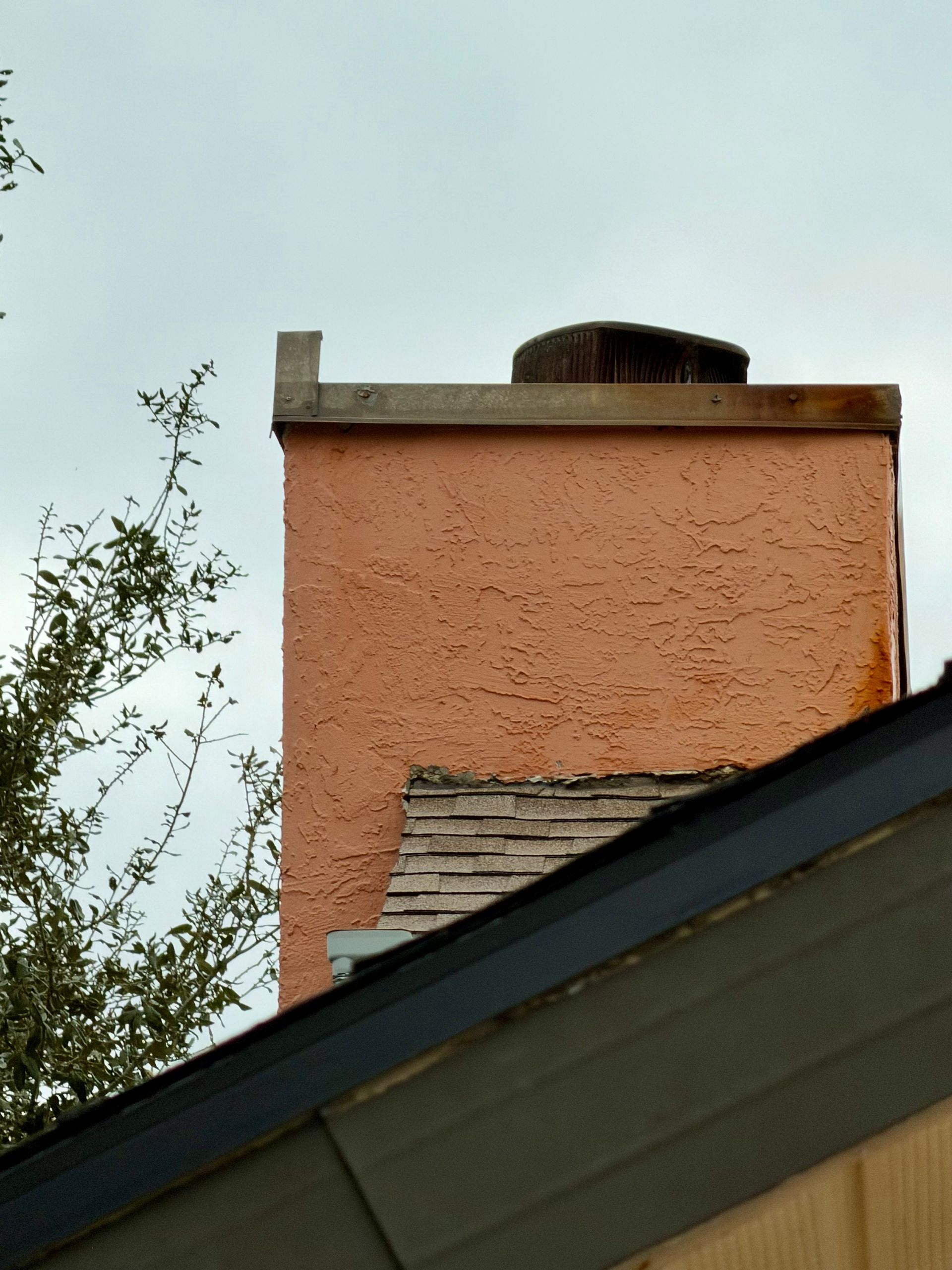 Orange stucco chimney with a metal cap above a sloped roof against a cloudy sky