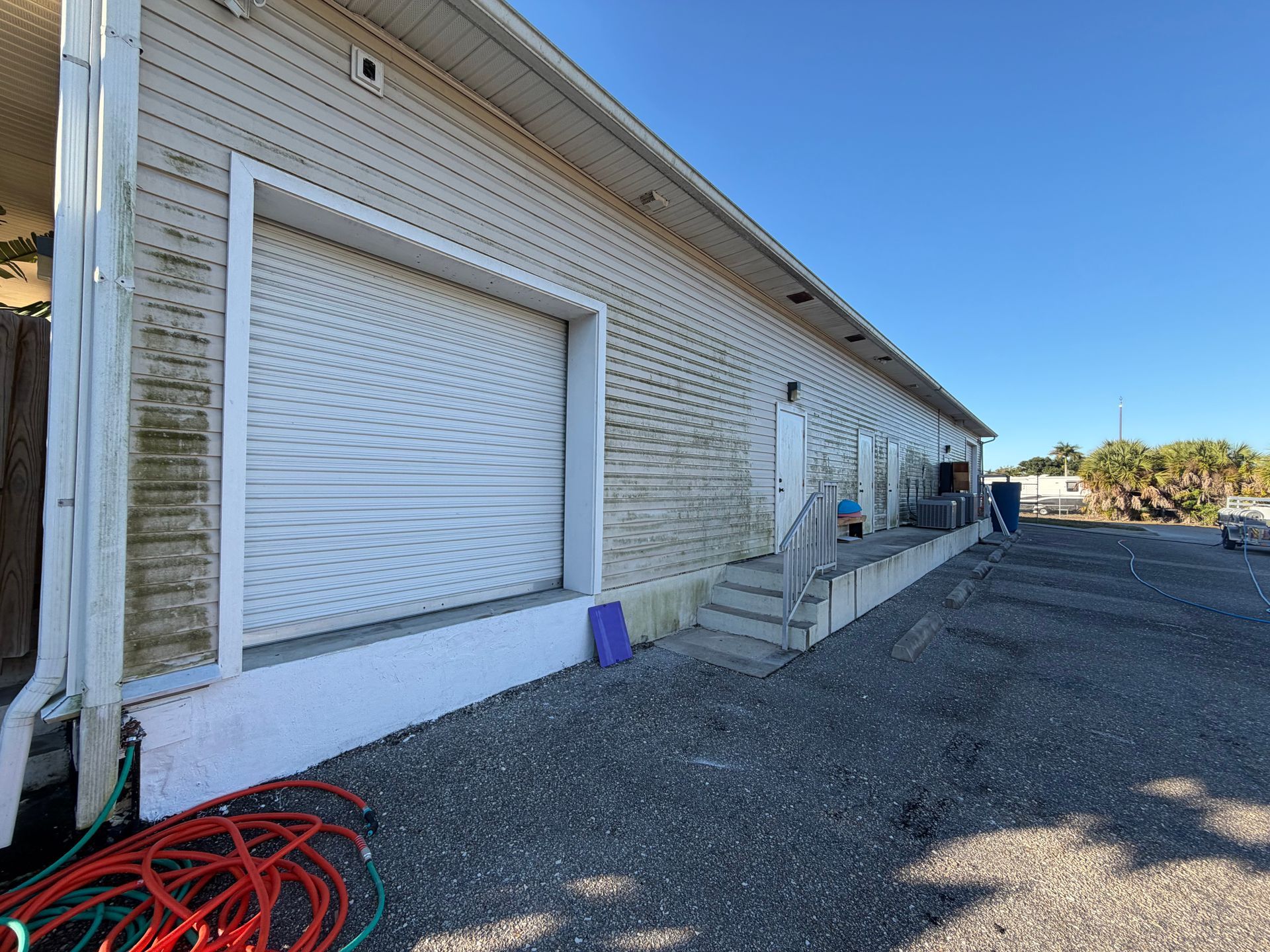White building with closed shuttered windows, steps, gravel lot, and coiled red hose under a blue sky