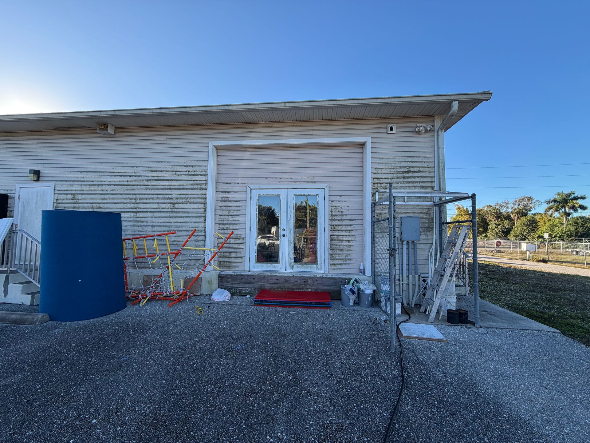 Exterior of a weathered building with a glass door, blue tank, and red kayak on gravel under a clear sky