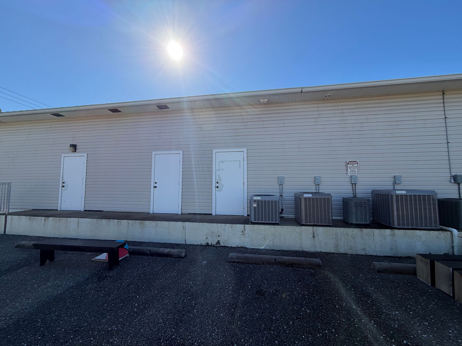Industrial building exterior with three white doors, stacked pallets, and bright sun overhead.