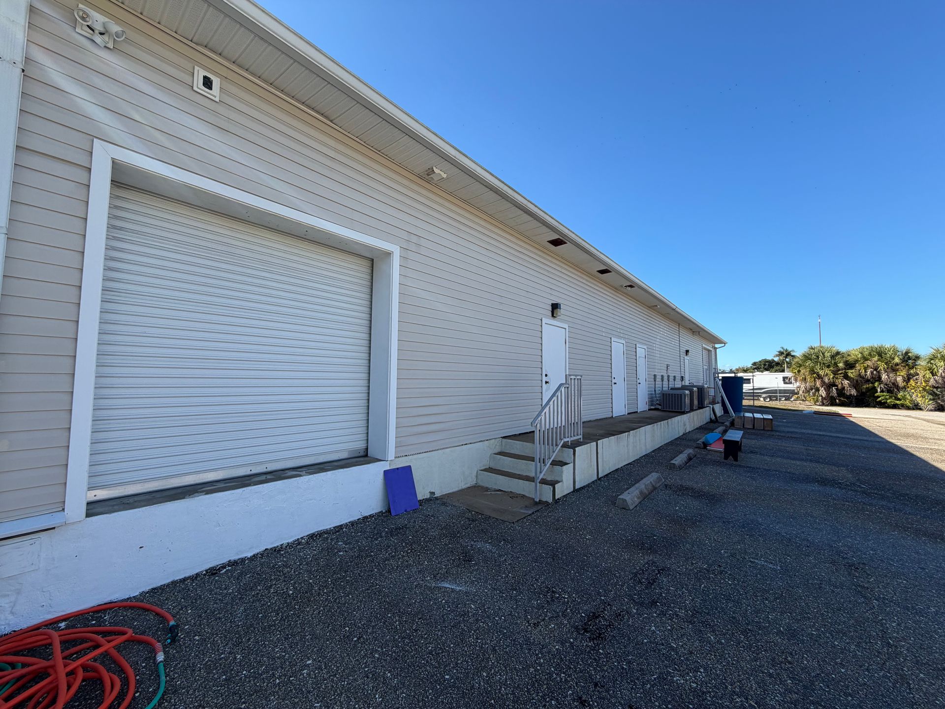 White industrial building with a raised loading dock, closed garage door, and gravel lot under a blue sky