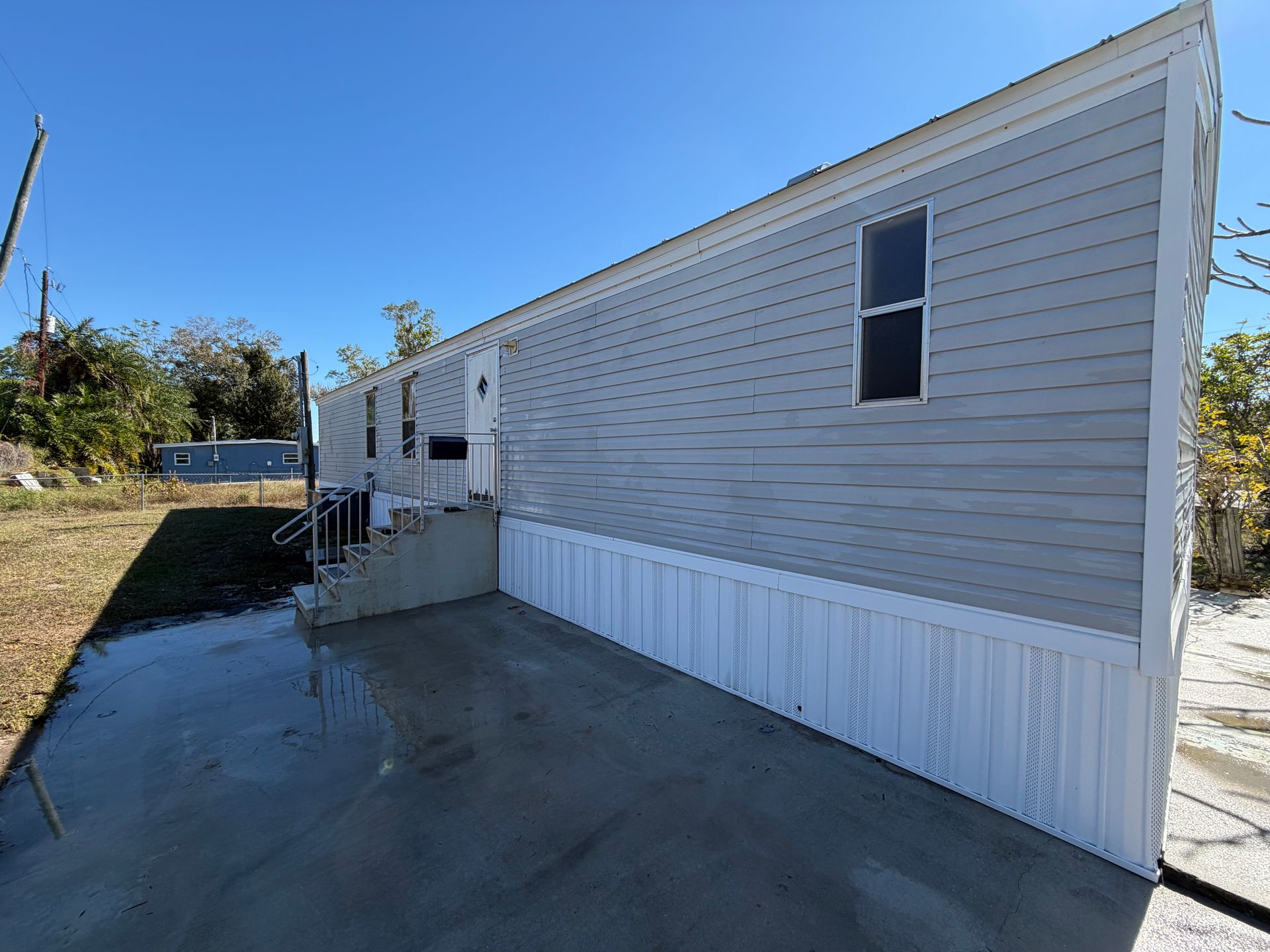 Gray mobile home exterior with a blue sky. Concrete patio and steps leading to the door.