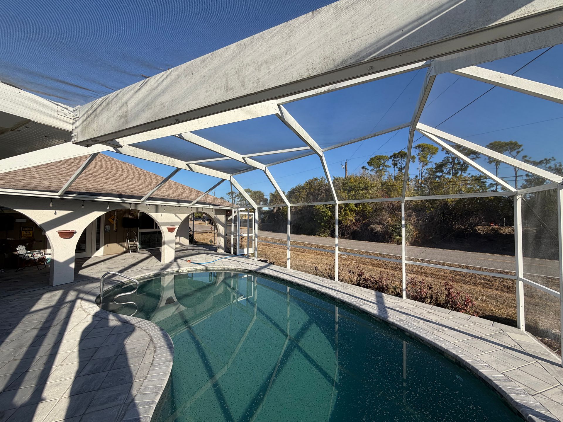 Screened pool enclosure over a turquoise backyard pool beside a white house under a blue sky