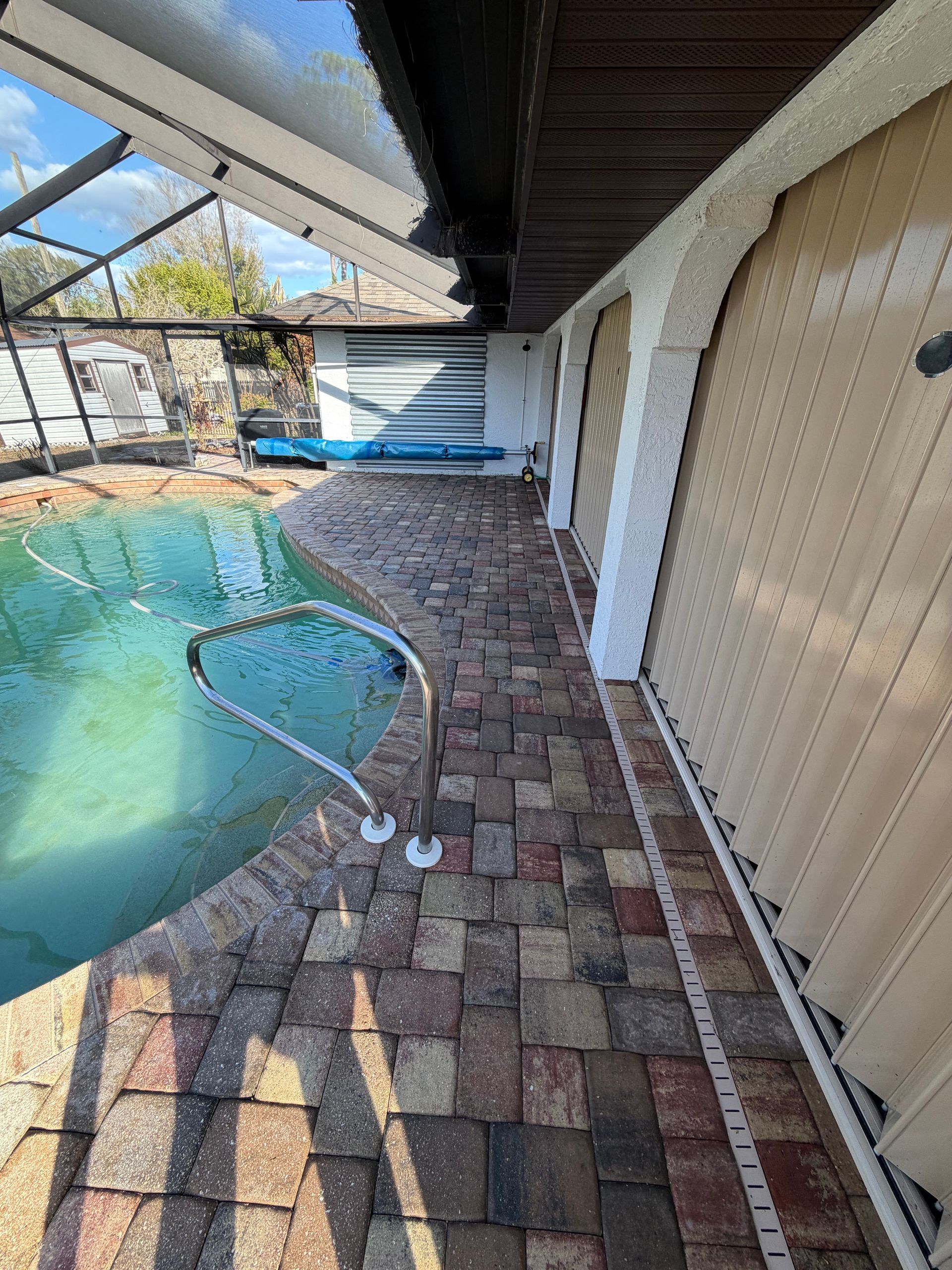 Poolside patio with brick pavers, a blue-tiled pool, and a metal hose or pole near a screened enclosure