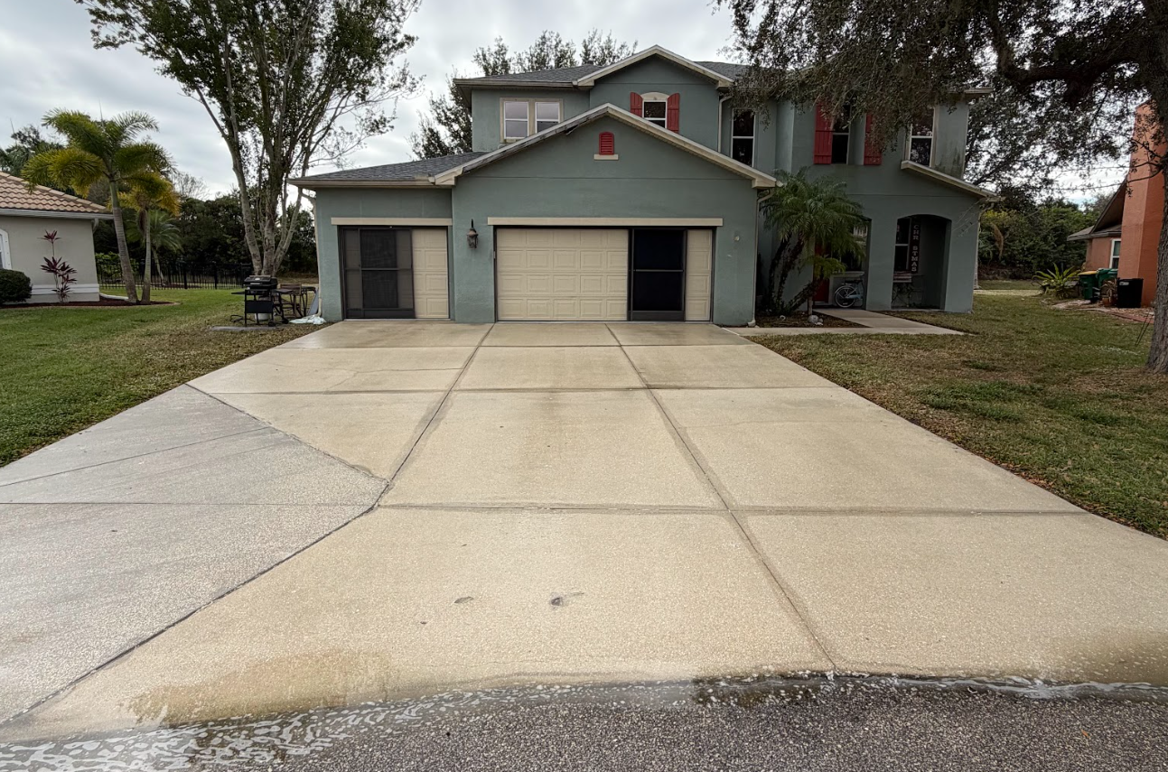 Driveway paved with textured stones, flanked by green grass and a silver SUV parked in front of a house. Palm tree shadow.