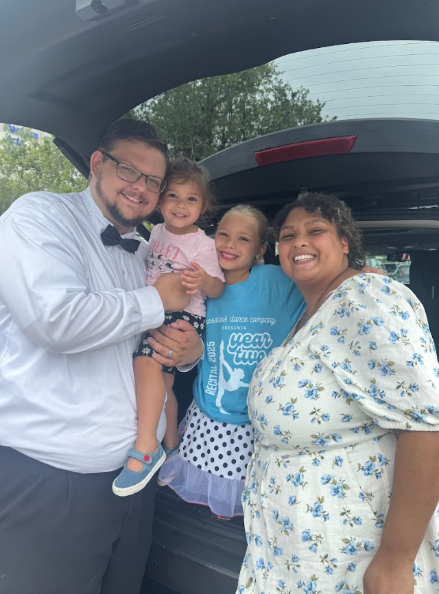 Family posing in a car trunk; man in tux holds a child, two girls wear dance attire, smiling.