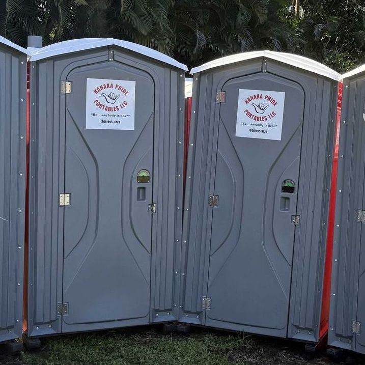 Two gray portable toilets with white roofs stand in a grassy area. Each has a sign on the door with text in Spanish.