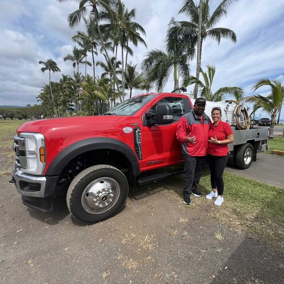 Two people stand next to a red Ford truck with a flatbed. They are smiling and wearing matching red shirts.
