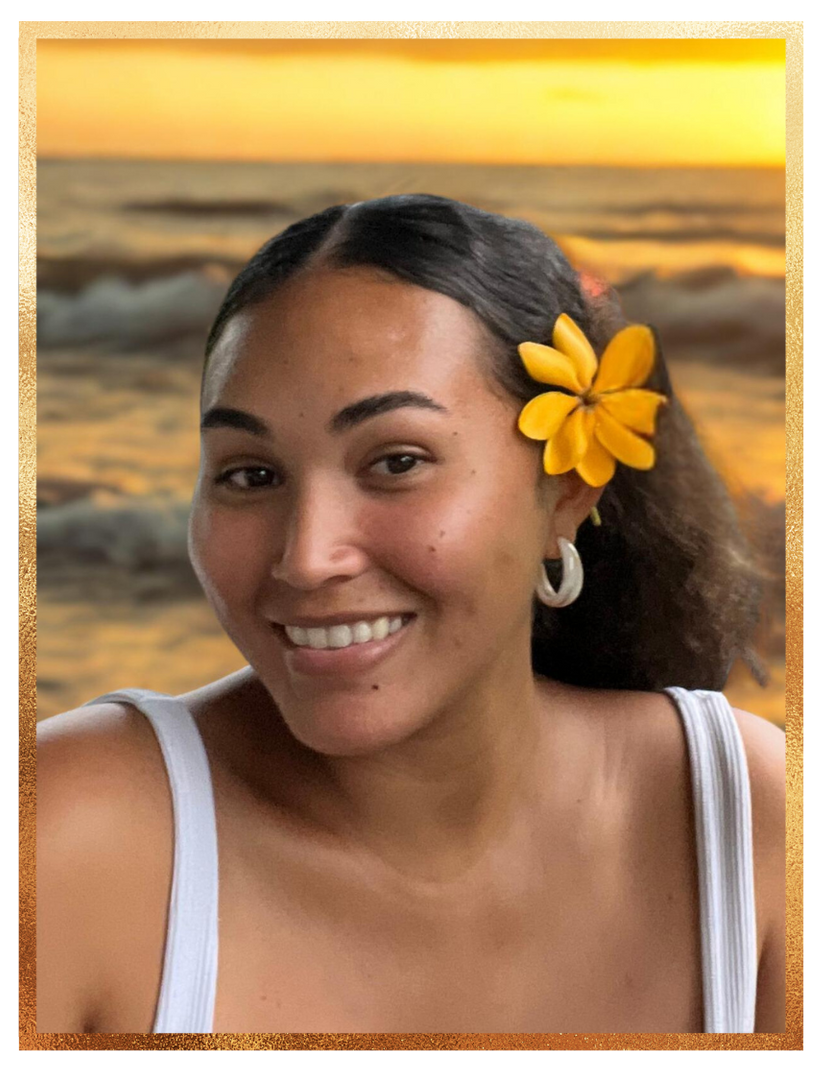 Woman smiling, wearing a yellow flower and white top, with ocean sunset in background.