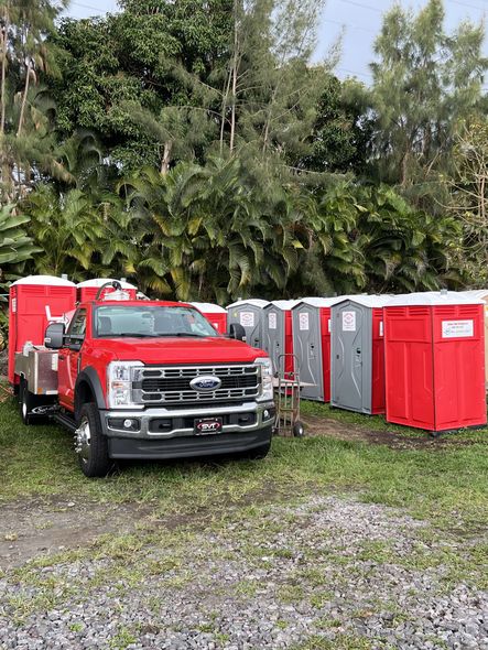 A red truck parked next to a row of portable toilets in a grassy area, with trees in the background.