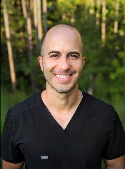 A bald man wearing a black scrub top is smiling for the camera.