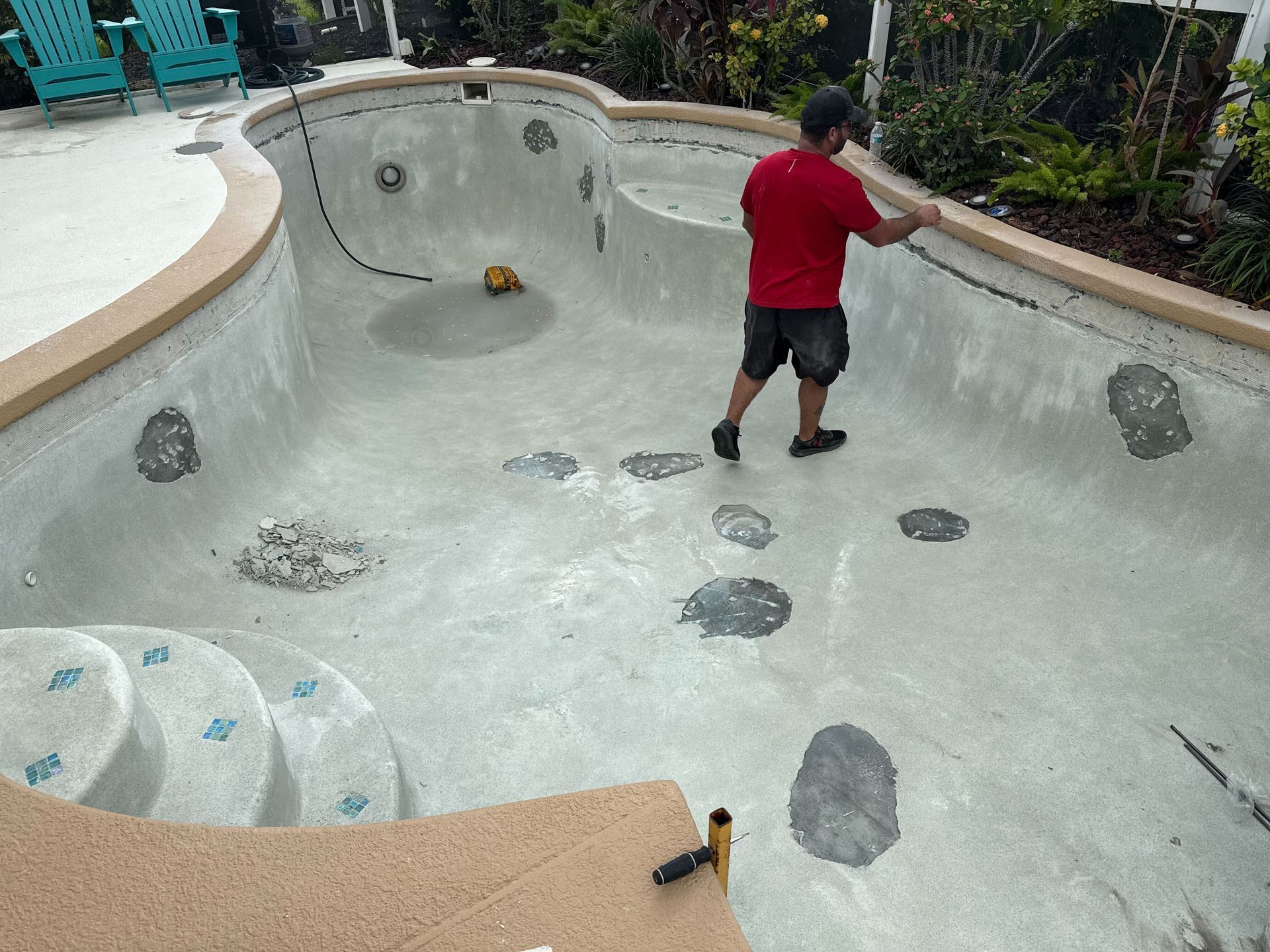 Man in red shirt working inside empty, concrete swimming pool with tan trim.