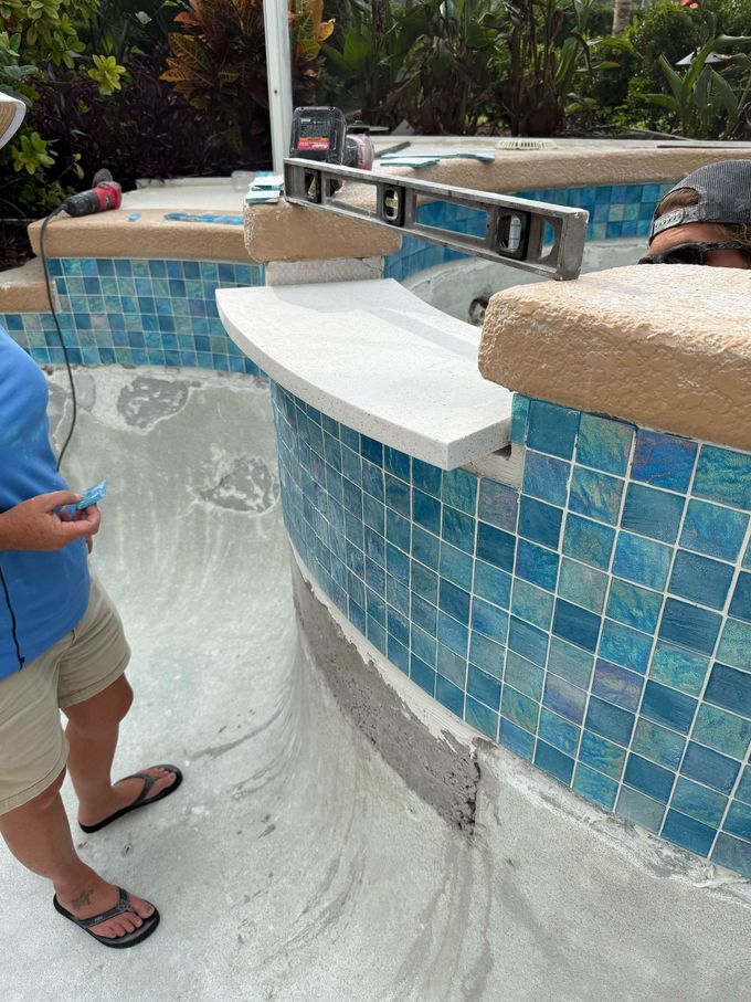 Person installing coping on a curved pool edge, checking level. Blue and tan tiles.