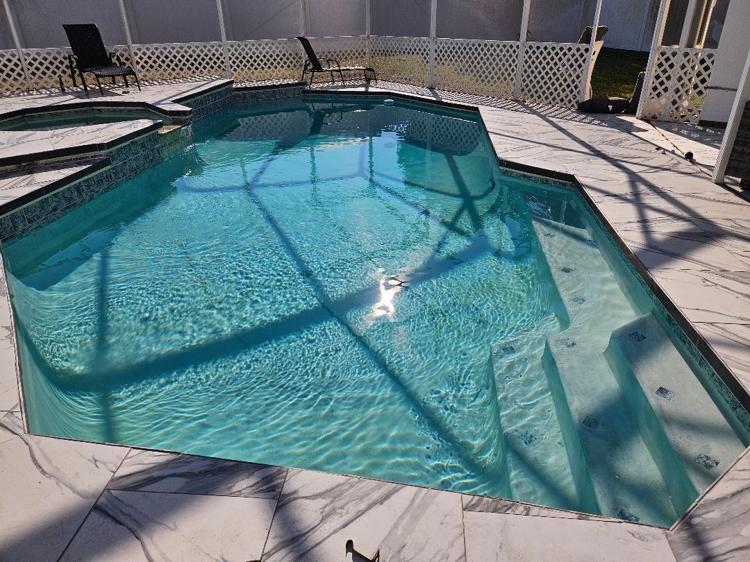 A swimming pool with clear blue water and steps, surrounded by white tile and a fence.