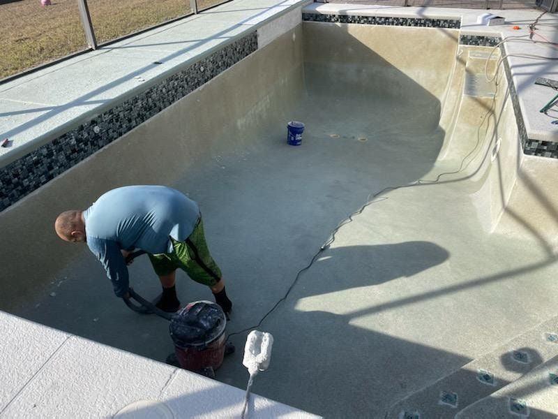 Person cleaning an empty swimming pool. Pool has light-colored walls, dark rocks around the edge, and steps. Sunny outdoors.