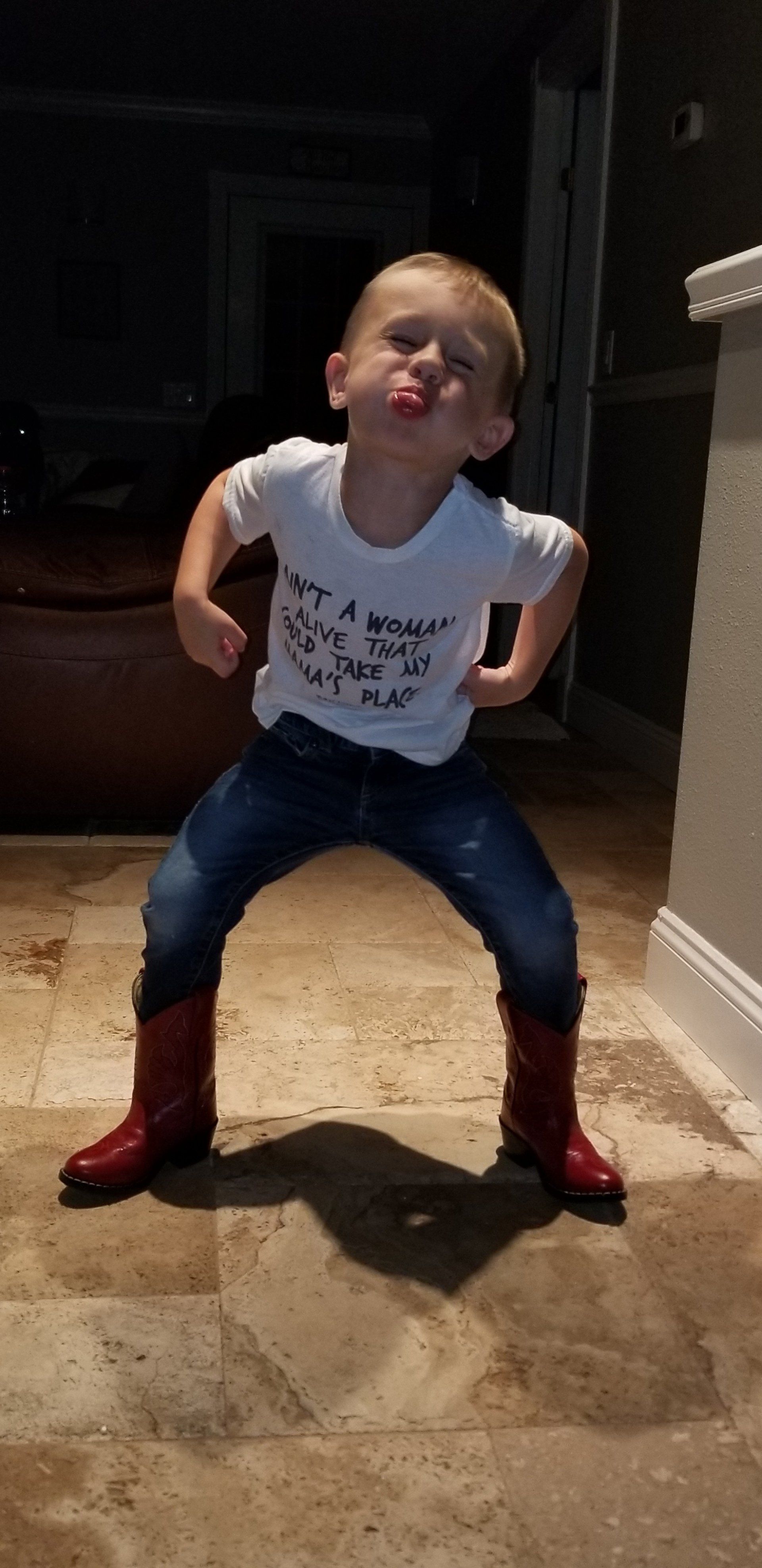 A little boy wearing cowboy boots and a white shirt is making a funny face.
