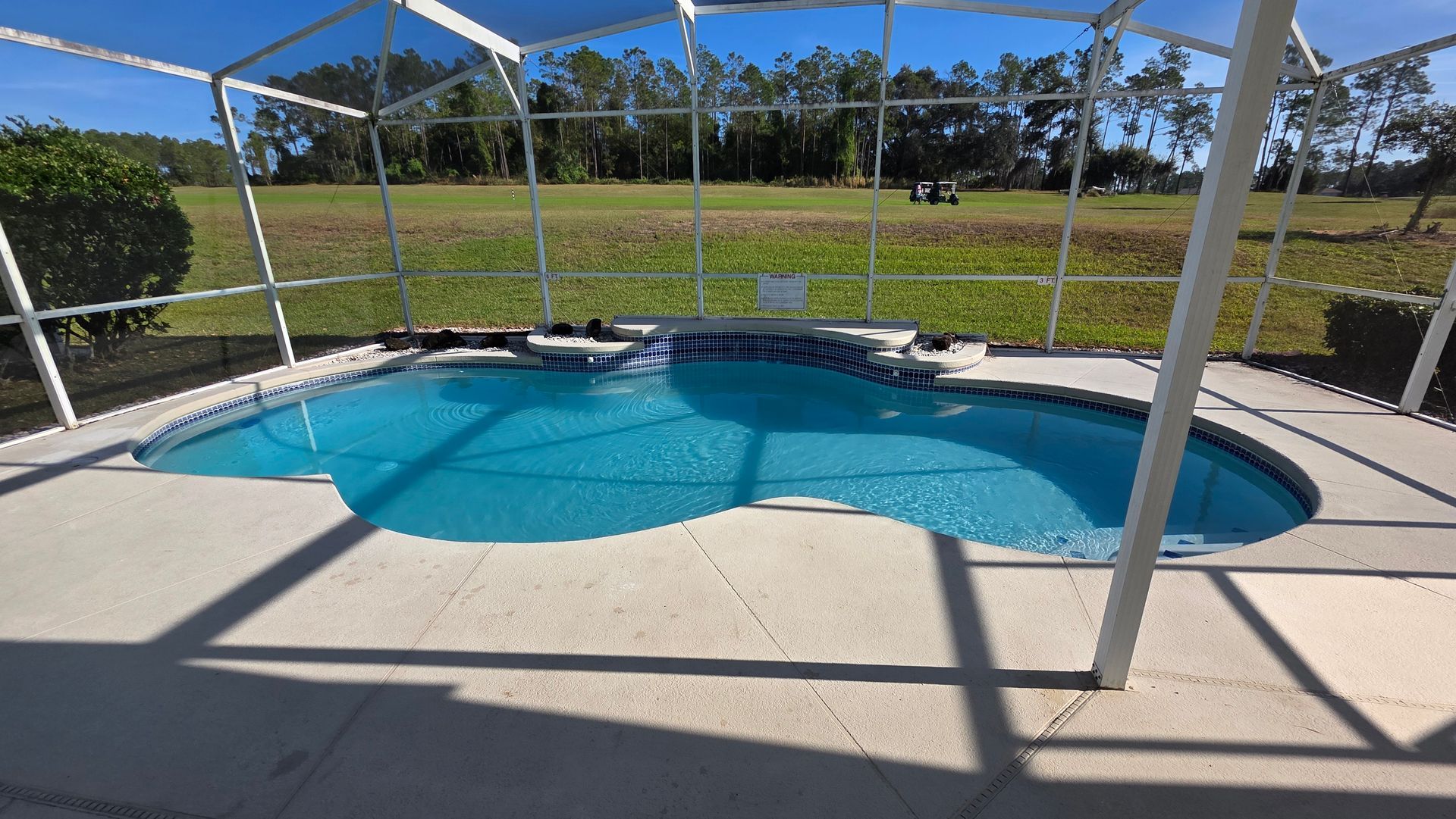 Swimming pool enclosed by a screen, with a clear view of a green field and trees in the distance.